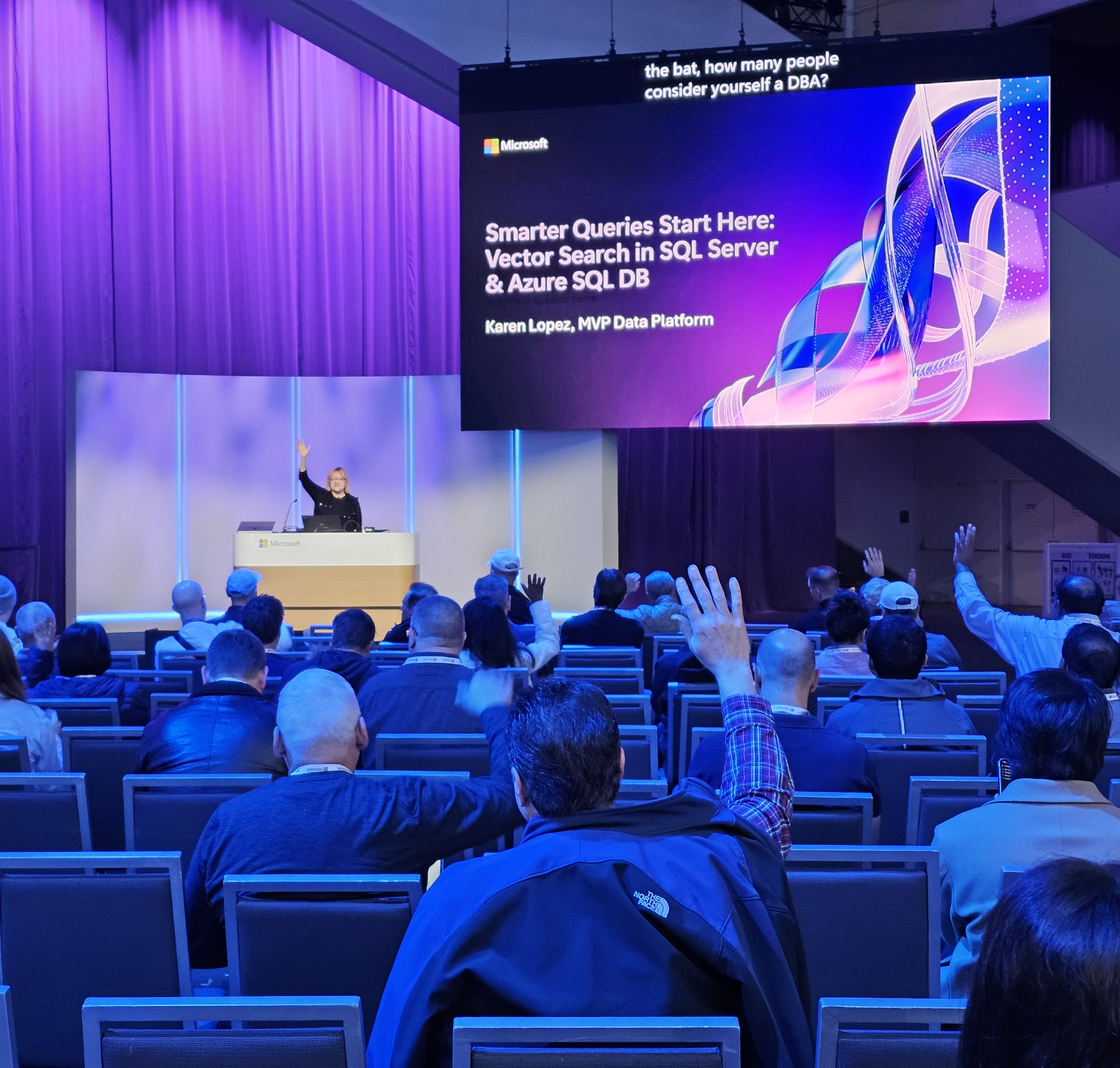 Karen speaking on a stage. Lighting is purple and blue. Audience members are raising their hands.